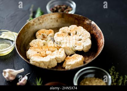 Blumenkohlsteak Kochen. Roher Blumenkohl, mit Gewürzen bestreut, liegt in einer Bratpfanne. Olivenöl, Kräuter, verschiedene Gewürze in der Nähe. Dunkler Hintergrund. Stockfoto