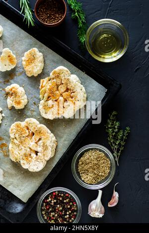 Blumenkohlsteak Kochen. Roher Blumenkohl, mit Gewürzen bestreut, liegt auf einem Backblech. Olivenöl, Kräuter, verschiedene Gewürze in der Nähe. Dunkler Hintergrund. Stockfoto