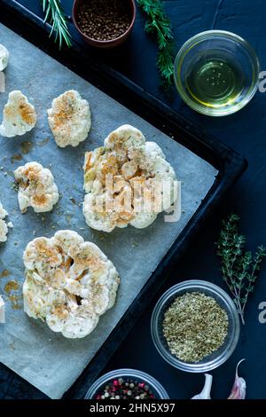 Blumenkohlsteak Kochen. Roher Blumenkohl, mit Gewürzen bestreut, liegt auf einem Backblech. Olivenöl, Kräuter, verschiedene Gewürze in der Nähe. Dunkler Hintergrund. Zinn Stockfoto