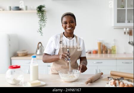 Lächelnde, hübsche, tausendjährige afroamerikanische Küchenchefin in Schürze, die Teig in einer hellen, minimalistischen Kücheneinrichtung zubereitet Stockfoto
