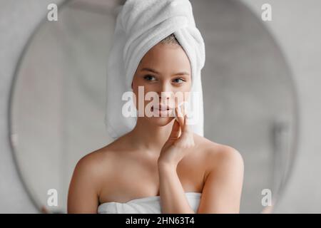 Schönheitsroutine. Hübsche junge Frau, die ihr Gesicht zu Hause mit Wattepad putzt und nach dem Bad mit einem Handtuch auf dem Kopf steht Stockfoto