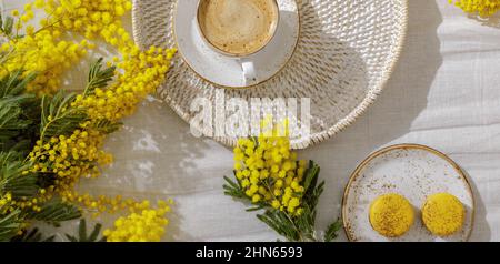 Zweige gelber Mimosenblüten auf weißen Leinenbettlaken, Tasse oder Kaffee, französische gelbe Makronen. Gruß zum Internationalen Frauentag am 8t. März Stockfoto