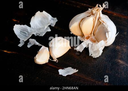 Einen Knoblauchkopf auf einem Holztisch schälen und schneiden: Knoblauchkopf mit Schale und Nelken auf dunklem Hintergrund Stockfoto