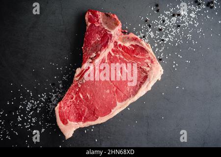 Rohes T-Bone Steak mit koscheres Salz und schwarzen Pfefferkörnern: Ein rohes Rindersteak mit Knochen und Salz und Pfeffer auf dunklem Grund Stockfoto