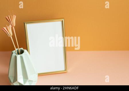 Leerer Fotorahmen mit Vase mit trockenen Blumen auf rosafarbenem Tisch. Orangefarbener Wandhintergrund. Kopierraum Stockfoto
