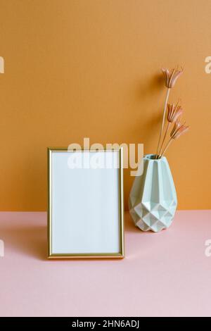 Leerer Fotorahmen mit Vase mit trockenen Blumen auf rosafarbenem Tisch. Orangefarbener Wandhintergrund. Kopierraum Stockfoto