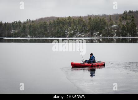 Teenager, der im Winter in einem Fischerkajak auf einem eisigen See sitzt. Stockfoto