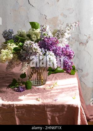 Vase mit frischen bunten Fliederblumen auf dem Leinentischtuch aus Leinenstoff mit einem sonnigen Tageslicht, Postkarten-Konzept für Frühling und Sommerferien Stockfoto