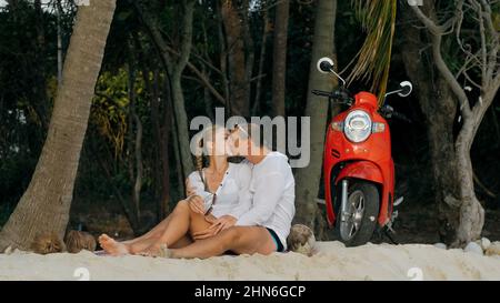 Motorroller Road Trip. Schönes Paar auf rotem Motorrad in weißen Kleidern am Sandstrand. Gerade verheiratete Menschen küssen Umarmungen, die in der Nähe der tropischen Palmen, dem Meer, spazieren gehen. Hochzeitsreise am Meer. Motorradmiete. Stockfoto