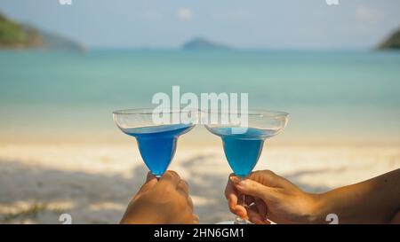 Das Liebespaar hält ein Glas blauen curacao Cocktail auf dem Meer. Nahaufnahme. Mann und Frau trinken Alkohol am Sandstrand im Schatten des azurblauen Ozeans einer tropischen Insel. Feiern Sie den Sommerurlaub. Stockfoto