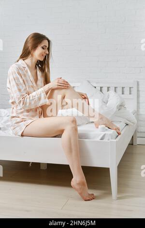 Junge, fröhliche Frau im Pyjama sitzt auf dem Bett und gibt Körperlotion auf das Bein. Frau trägt nach dem Epilieren oder Enthaarung eine Bademantel-Reinigungslotion. Stockfoto