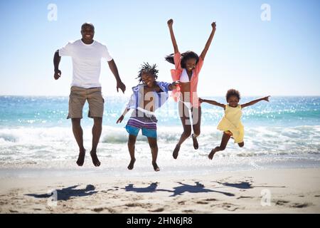 Woohoo Urlaub rockt. Eine aufgeregte afroamerikanische Familie, die am Strand mit einem wunderschönen Meer im Hintergrund in die Luft springt. Stockfoto