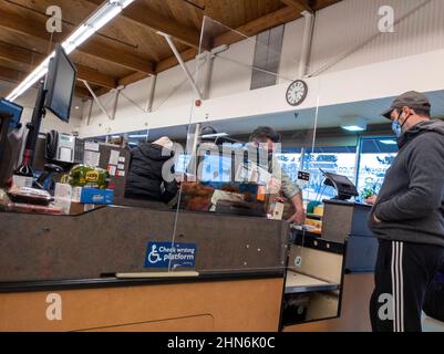 Kirkland, WA USA - ca. Dezember 2021: Ansicht eines männlichen Kassierers hinter einem Plexiglas-Trennwand an einem Kassenschalter in einem PCC-Lebensmittelgeschäft während des Cor Stockfoto