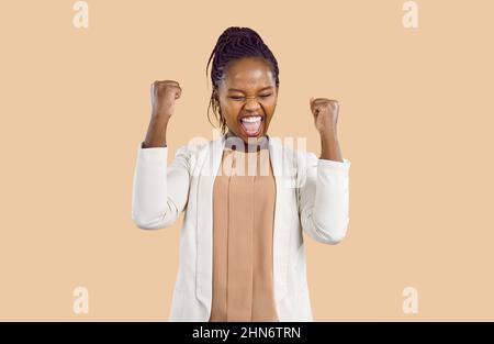 Triumphende glückliche und zufriedene junge afroamerikanische Frau freut sich aufrichtig auf beigen Hintergrund. Stockfoto