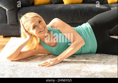 Fröhliche und aktive ältere Frau, die zu Hause Stretching macht. Großmutter in Sportkleidung, die sich aufwärmt und morgendliche Übungen macht Stockfoto