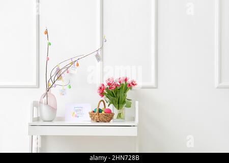 Schöne Osterkomposition mit Korb, bemalten Eiern, Blumen und Grußkarte auf dem Tisch in der Nähe der weißen Wand Stockfoto