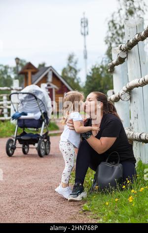 Mutter küsst und umarmt ihre kleine Tochter im Park. Menschen, die mit Kinderwagen laufen, Frauen mit zwei Kindern Stockfoto