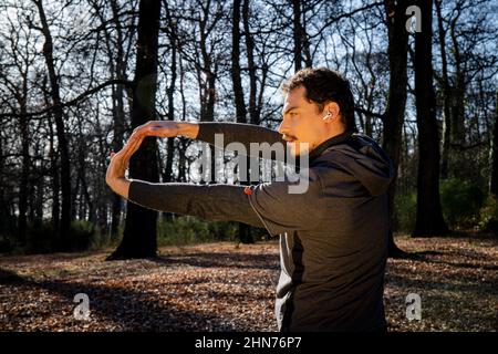 Läufer macht Stretching im Wald. Junger Mann mit blauen Augen und Schnurrbart trainiert im Park. Stockfoto