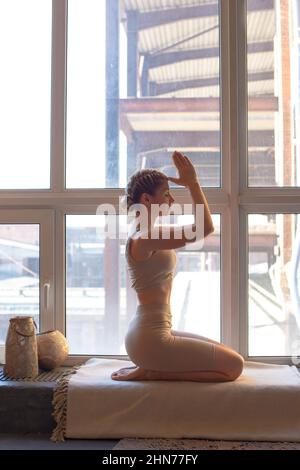 Eine schlanke und schöne Frau in einem beigen Oberteil und Leggings sitzt an einem großen Fenster in einer Yoga-Pose Stockfoto