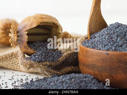 Nahaufnahme eines gebrochenen trockenen Mohn mit reifen Mohnblumen aus tiefem Winkel. Weißer Holztisch mit Holzschüssel voller Mohnsamen. Stockfoto