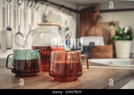 Teekannen aus Glas mit schwarzem Tee und zwei Tassen Tee auf der Küchenarbeitsfläche in der Nähe Stockfoto