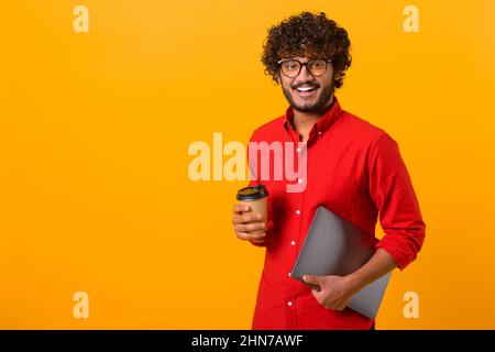 Bild von einem gutaussehenden, fröhlich lächelnden indischen Mann, der die Hand ausstreckt, eine Tasse Kaffee zum Mitnehmen hält, Kaffee oder Tee trinkt und auf orangefarbenem Hintergrund mit der Kamera lächelt Stockfoto