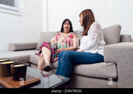 Behinderte kaukasische Erwachsene Frau zu Hause auf dem Wohnzimmer Sofa sitzen im Gespräch mit ihrer Mutter, häusliches Leben zwischen Mutter und Tochter mit Down-syndrom Stockfoto