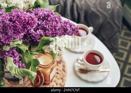 Bouquet von lila Flieder Blumen auf Couchtisch mit zwei Tassen Tee und Kerze in der häuslichen Einrichtung Stockfoto