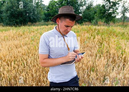 Junger Bauer. Porträt eines Bauern, der im goldenen Weizenfeld sitzt und auf dem Tablet scrollt. Junger Mann mit Cowboyhut auf dem Feld, der Weizenernte untersucht. Hafer Stockfoto
