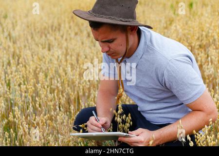 Digitale Farm. Porträt eines Bauern, der in einem goldenen Weizenfeld sitzt und auf einem Tablet schreibt. Junger Mann mit Cowboyhut auf dem Feld, der Weizenernte untersucht. Hafer Stockfoto