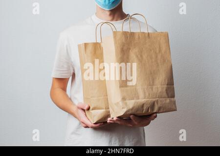 Junger Mann in schützender Gesichtsmaske, der Papiertüten in den Händen hält. Mann Kurier mit Einkaufstasche. Sichere Online-Lieferung vom Supermarkt nach Hause Stockfoto