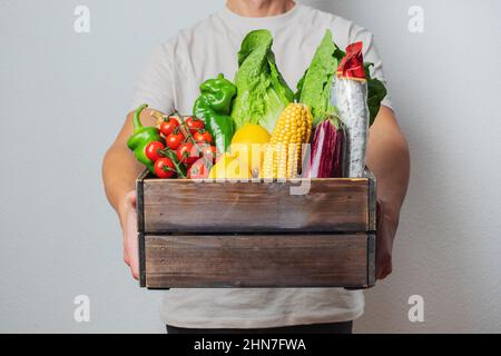 Lieferkonzept -Junge kaukasische Lieferer trägt Holzkiste mit Lebensmittelgemüse und Obst aus dem Laden. Isoliert auf grauem Studiohintergrund Stockfoto