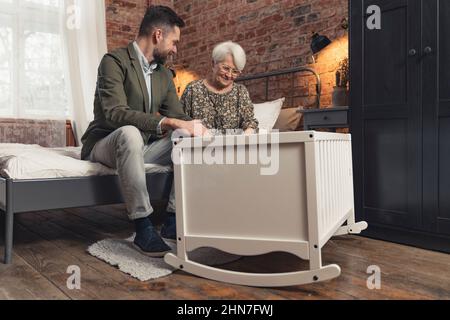 Stolze Großmutter und Novize Papa amüsiert sich über den Anblick eines neugeborenen Säuglings, der sich in einer Wiege legt. Hochwertige Fotos Stockfoto