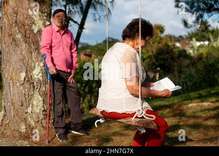 Ältere Frau liest ein Buch auf einer hölzernen Schaukel und älterer Mann im Hintergrund neben einem Baum. Stockfoto