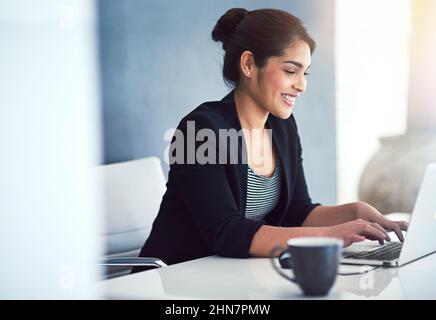 Sie arbeitet immer mit einem Lächeln im Gesicht. Eine kurze Aufnahme einer attraktiven jungen Geschäftsfrau, die in ihrem Büro an einem Laptop arbeitet. Stockfoto