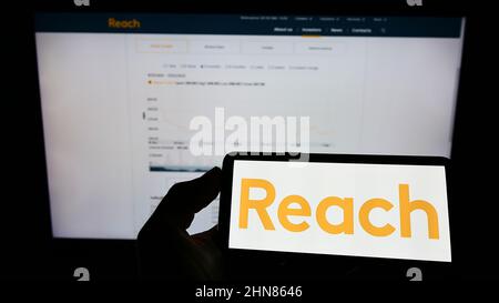 Person, die ein Mobiltelefon mit dem Logo des britischen Verlags Reach plc auf dem Bildschirm vor der Unternehmenswebseite hält. Konzentrieren Sie sich auf die Telefonanzeige. Stockfoto