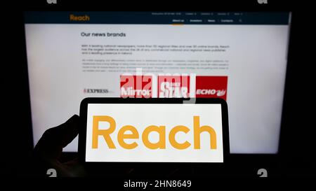 Person, die Mobiltelefon mit dem Logo des britischen Verlags REACH plc auf dem Bildschirm vor der Business-Webseite hält. Konzentrieren Sie sich auf die Telefonanzeige. Stockfoto