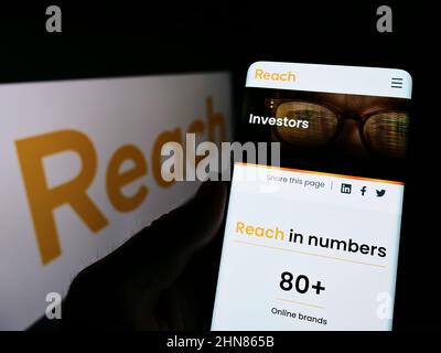 Person, die Mobiltelefon mit der Webseite des britischen Verlags Reach plc auf dem Bildschirm vor dem Logo hält. Konzentrieren Sie sich auf die Mitte des Telefondisplays. Stockfoto