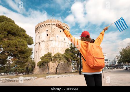 Ein Studentenmädchen mit Rucksack und griechischer Flagge auf dem Hintergrund eines Weißen Turms in Thessaloniki. Das Konzept der Staatsbürgerschaft und das Erlernen der griechischen Sprache Stockfoto