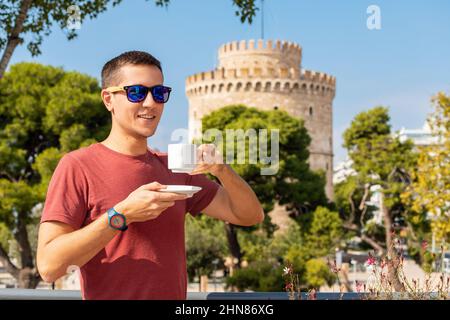 Ein fröhlich lächelnder junger Mann trinkt Kaffee aus einer Tasse vor dem Hintergrund des berühmten Weißen Turms in der Stadt Thessaloniki in Griechenland. Stockfoto