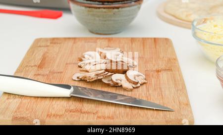 Crimini-Pilzscheiben auf Holzbrett auf Küchentisch aus nächster Nähe. Pizza mit Mozzarella und Pilzen Schritt für Schritt Rezept Stockfoto