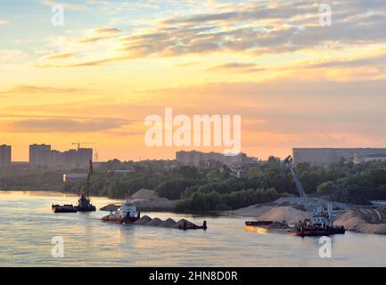 Nowosibirsk, Sibirien, Russland - 08.20.2020: Ein Schlepper mit einem beladenen Lastkahn fährt zu einem schwimmenden Kran, Sandhaufen am Ufer, die Häuser eines großen Kr Stockfoto