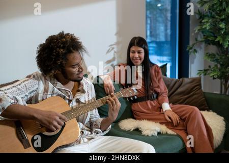 Afrikanischer junger Mann, der Gitarre spielt und ein Lied singt, während er mit seiner Freundin im Zimmer auf dem Sofa sitzt Stockfoto