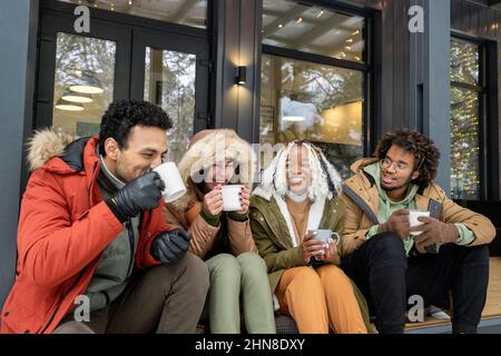 Gruppe von glücklichen Freunden sitzen auf der Veranda des Landhauses und genießen den Kaffee zusammen während des Wintermorgens im Freien Stockfoto