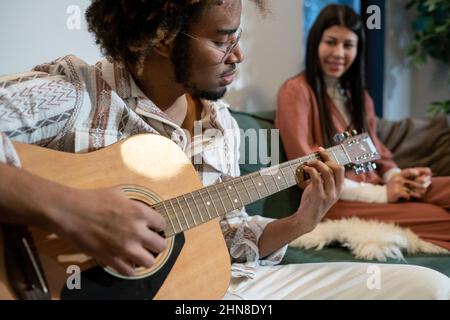 Afrikanischer junger Mann in einer Brille, der Gitarre spielt und ein Lied singt, während er auf dem Sofa sitzt, mit einer Frau im Hintergrund, die ihm zuhört Stockfoto