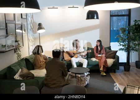 Eine Gruppe junger Menschen, die auf dem Sofa sitzen und Lieder zur Gitarre singen, verbringen zu Hause Zeit miteinander Stockfoto