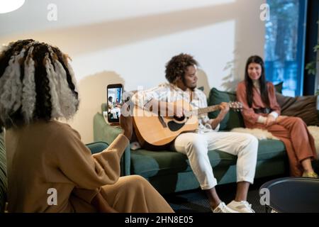 Rückansicht einer afrikanischen Frau, die ein Video von ihrem Mobiltelefon eines Mannes macht, der während ihrer Heimparty Gitarre spielt Stockfoto