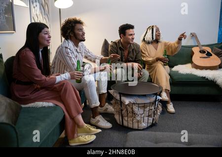 Eine Gruppe junger Freunde, die auf dem Sofa sitzen und Bier trinken, während sie sich gemeinsam im Zimmer zu Hause ein Sportspiel ansehen Stockfoto