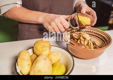 Hände des Kochs schneiden Schale von rohen Kartoffeln mit Küchenmesser zum Schälen von Gemüse, Schäler, legen Lebensmittelabfälle in separaten Behälter für die weitere Verarbeitung. Stockfoto