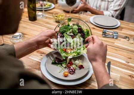 Nahaufnahme eines Mannes, der am Tisch sitzt und während der Abendparty Gemüsesalat aus der Schüssel auf seinen Teller legt Stockfoto